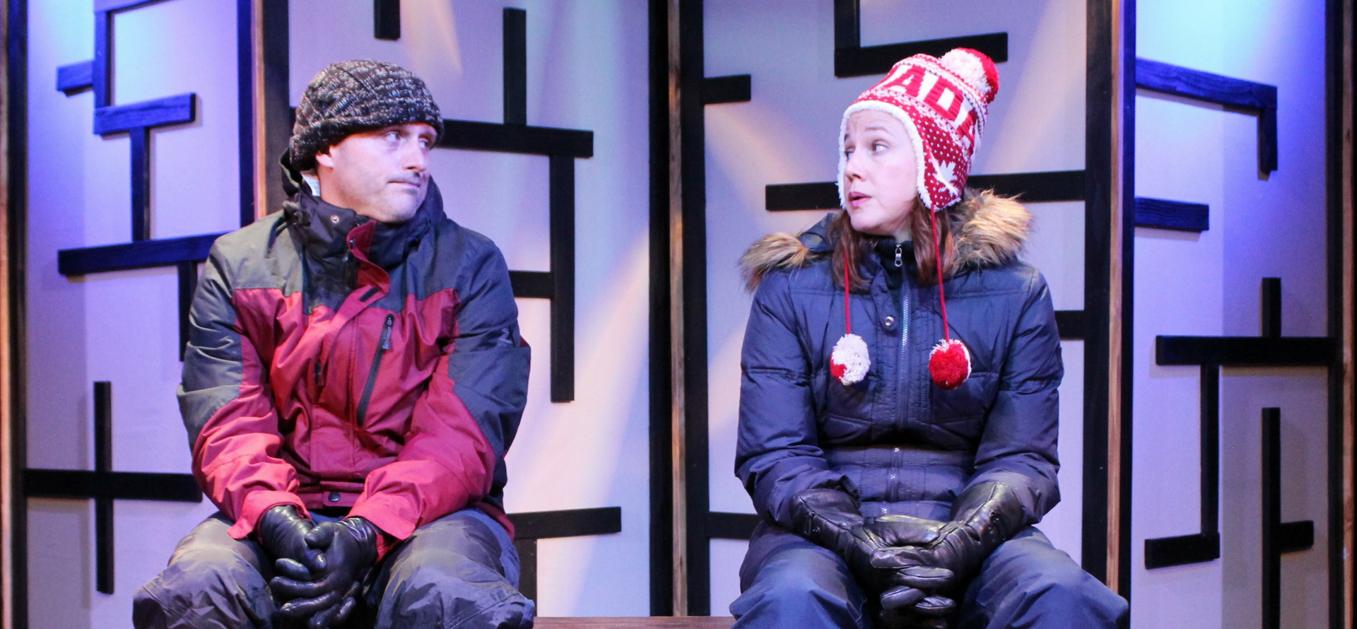 A man and woman on stage in play, sitting on chairs wearing snow gear