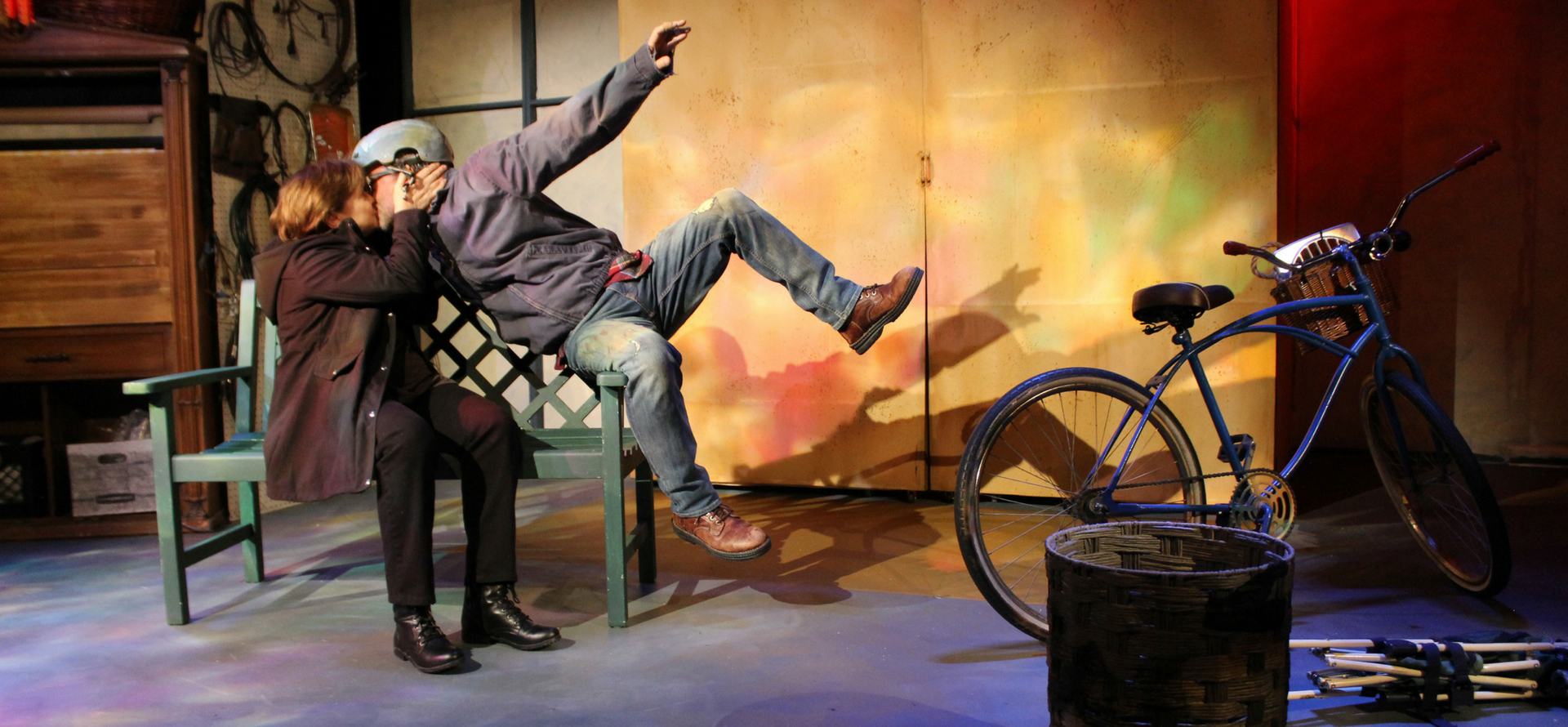 Man and woman on stage in play kissing; the man had just fallen off of a bike that is in the background