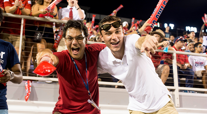 Two male students during an FAU football game smiling and pointing at the camera