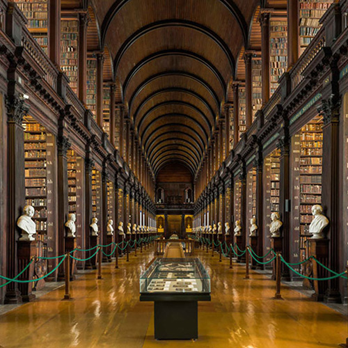 A long, symmetrical library hall lined with tall wooden bookshelves, statues, and a vaulted ceiling, with a central display case.