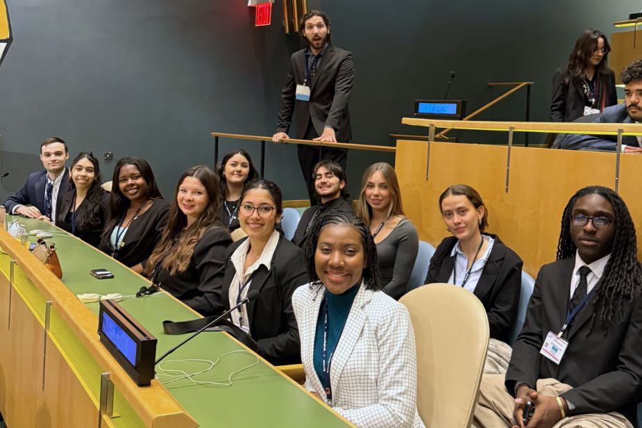 A large group of diverse students smiling sitting in 2 rows of chairs inside the United Nations, New York
