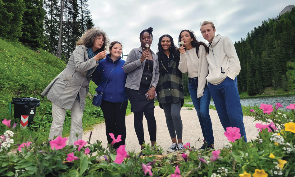 A group of students and a professor are blowing the seeds off dandelions while standing among the Dolomites mountains in Italy. There mountains in the backdrop are covered with grass and trees and brightly colored flowers are in the foreground of the image