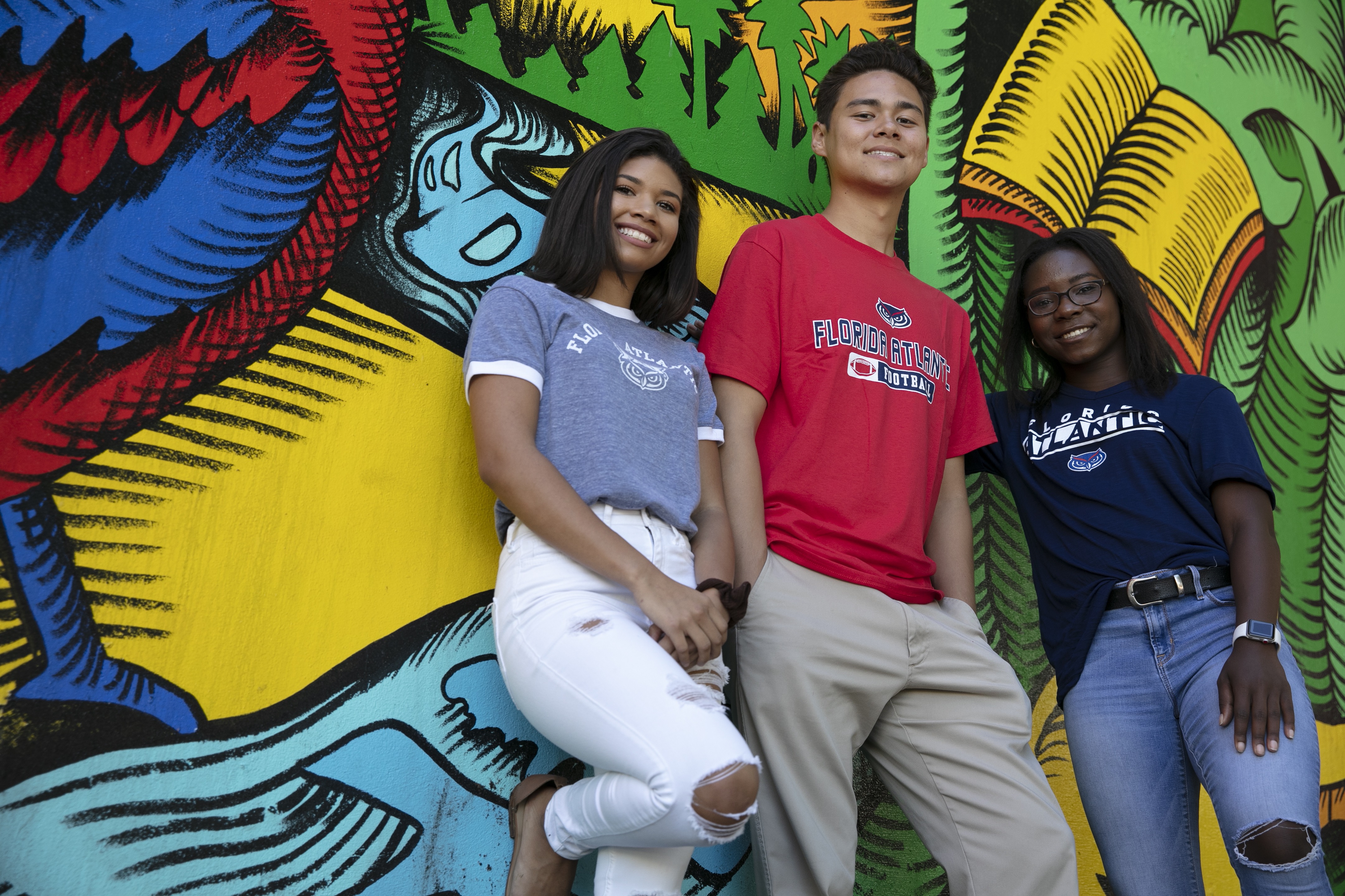 three students wearing FAU shirts stand in front of a mural while smiling at the camera