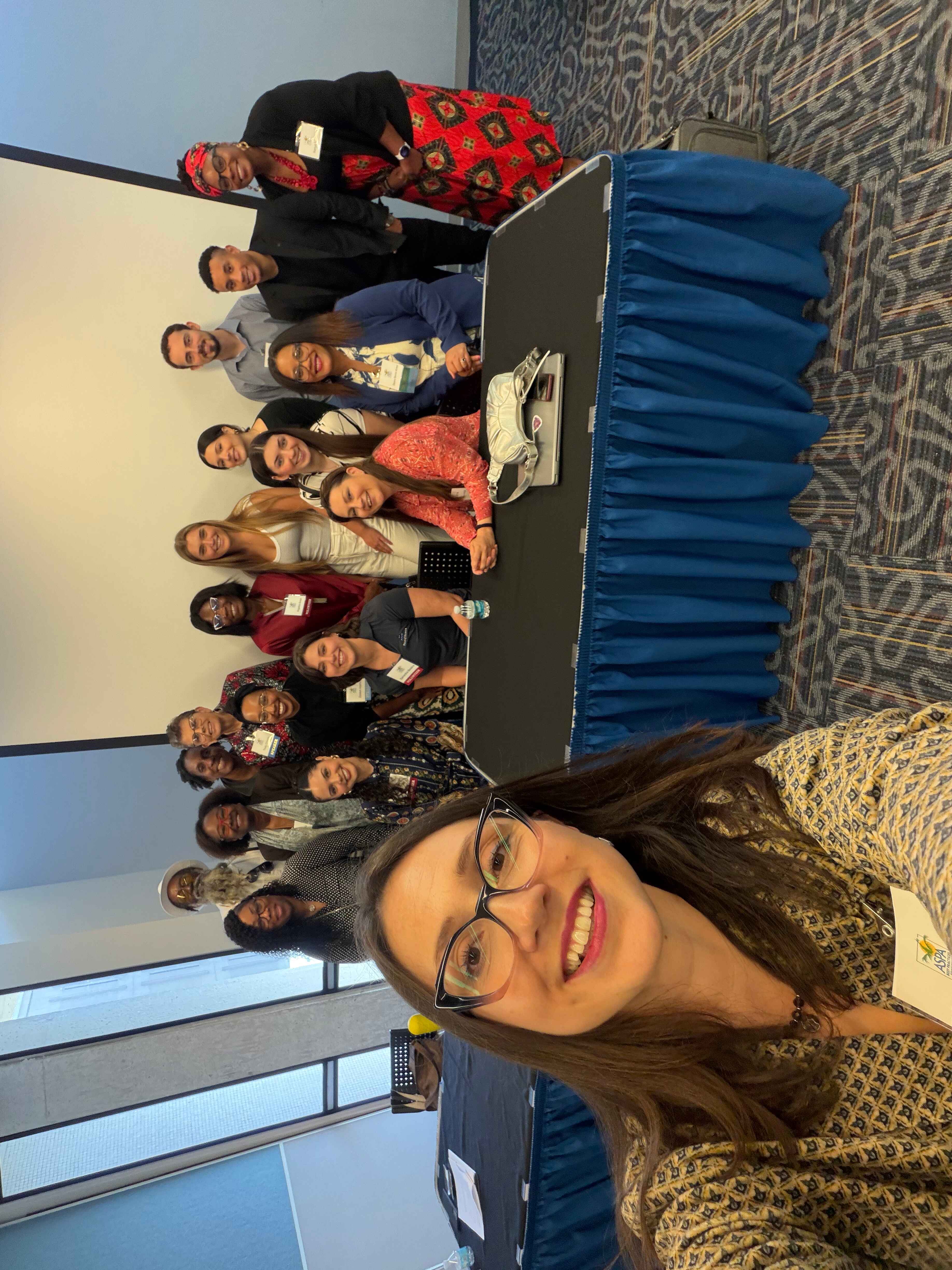 A large group poses for a group photo behind a conference table, smiling toward the camera. A woman in the foreground takes the selfie, and name badges and conference materials are visible on the table.