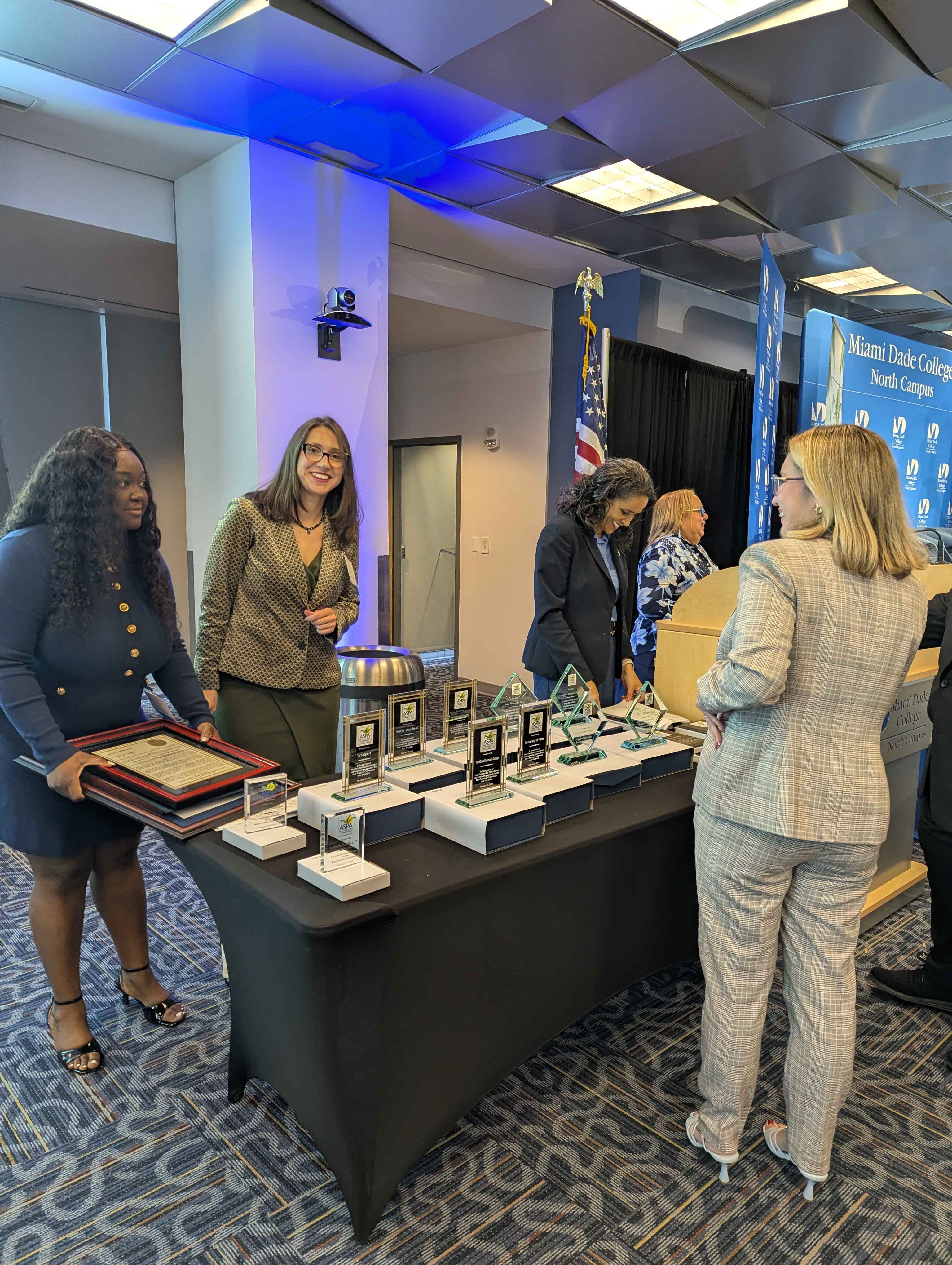 Several people stand behind a conference booth displaying awards, plaques, and informational materials. One attendee speaks with a booth representative while banners and signage for Miami Dade College appear in the background.