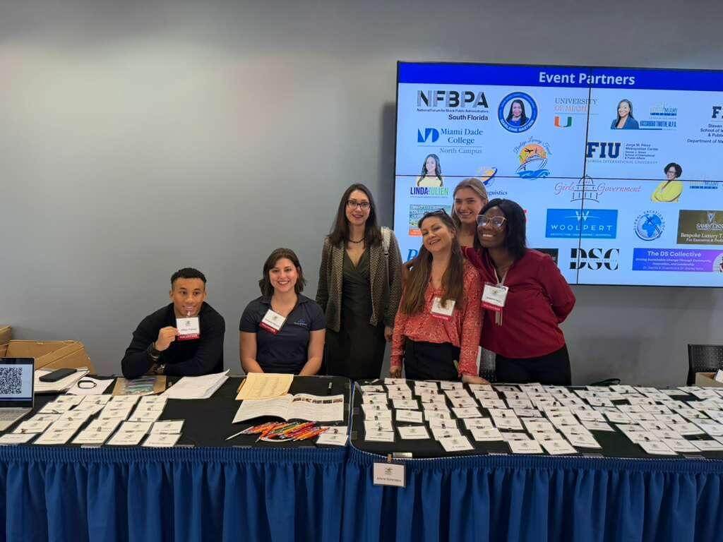 Six people stand and sit behind a registration table covered with name badges at a professional conference. A large screen behind them displays “Event Partners” along with multiple organizational logos.
