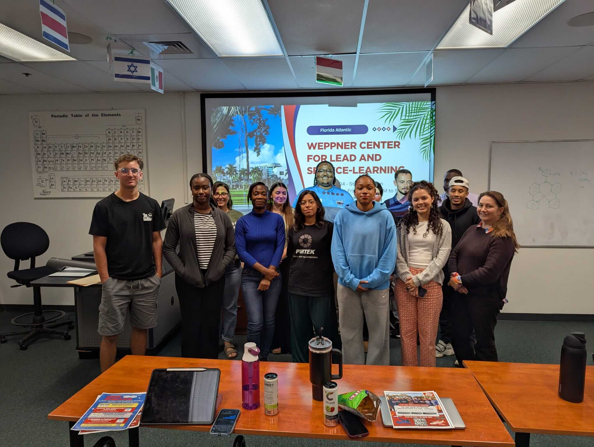 A group of students and facilitators stand together in a classroom in front of a projection screen that reads “WEPPNER CENTER FOR LEAD AND SERVICE-LEARNING.” Desks with notebooks and water bottles are in the foreground, and international flags hang from the ceiling.