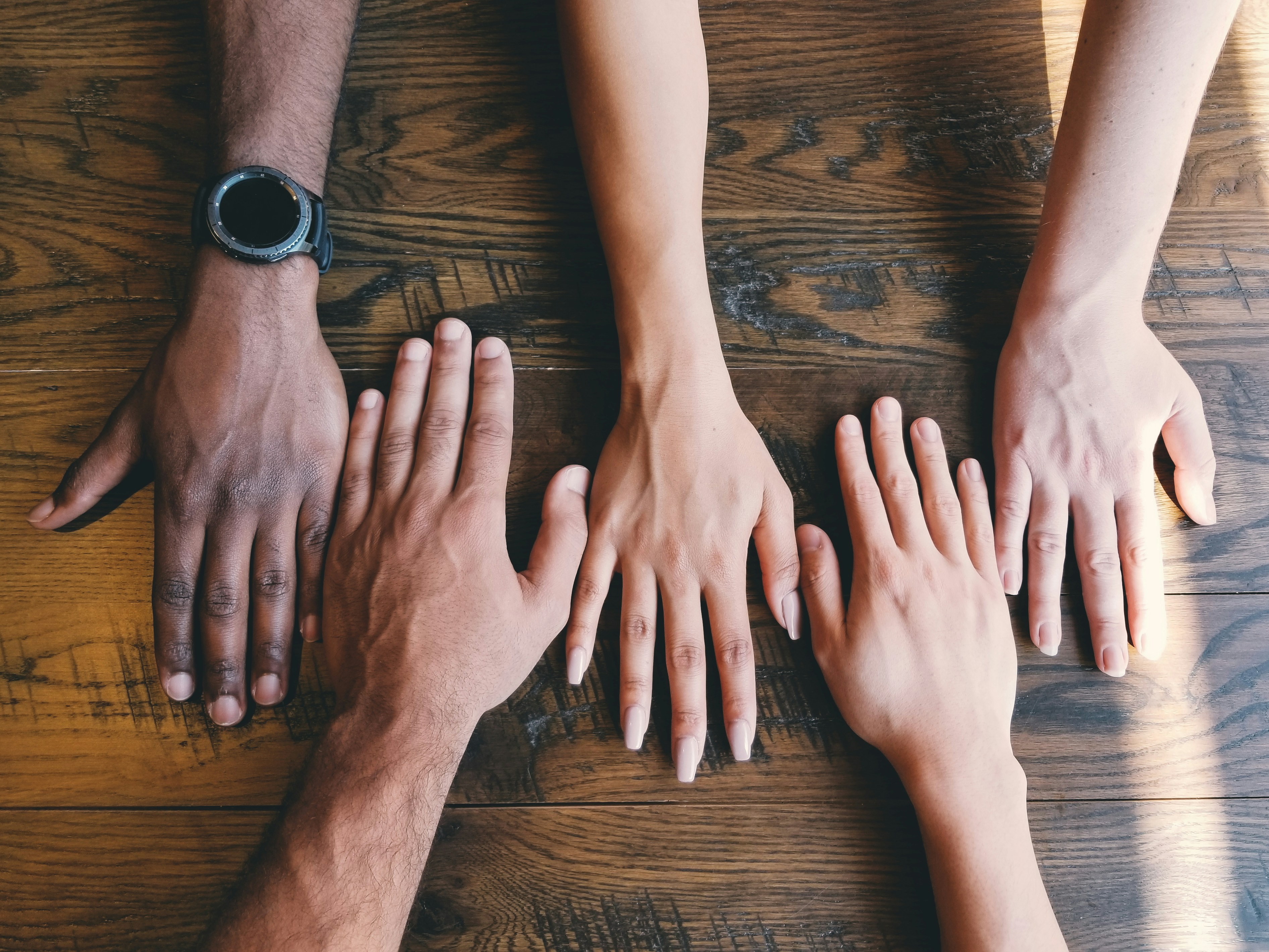 Photograph of five hands with different skin colors ranging from fair to dark on a wooden table.