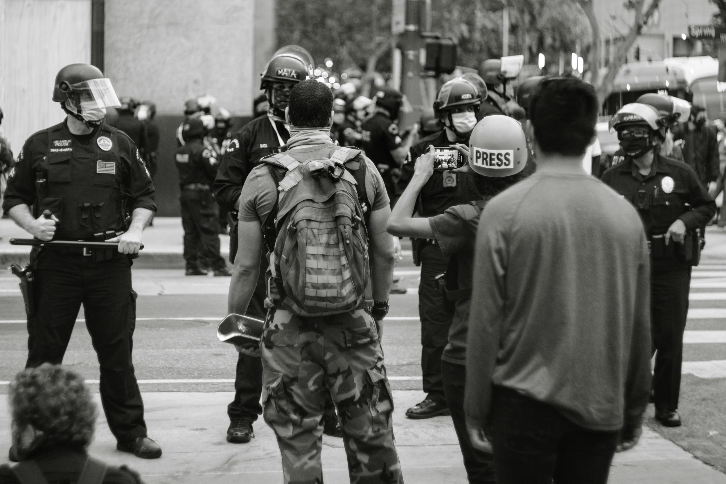 Black and white photograph showing a group of police officers in riot gear facing several civilians on a city street. Key elements include officers wearing helmets and holding batons, a person with a backpack holding a megaphone, and a press member filming the scene, highlighting a tense public protest or confrontation.
