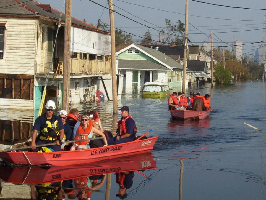 Photograph showing U.S. Coast Guard personnel in two red rescue boats navigating a flooded residential area with partially submerged houses and vehicles. The scene highlights emergency response efforts during a severe flood, with rescuers wearing life jackets and assisting residents.