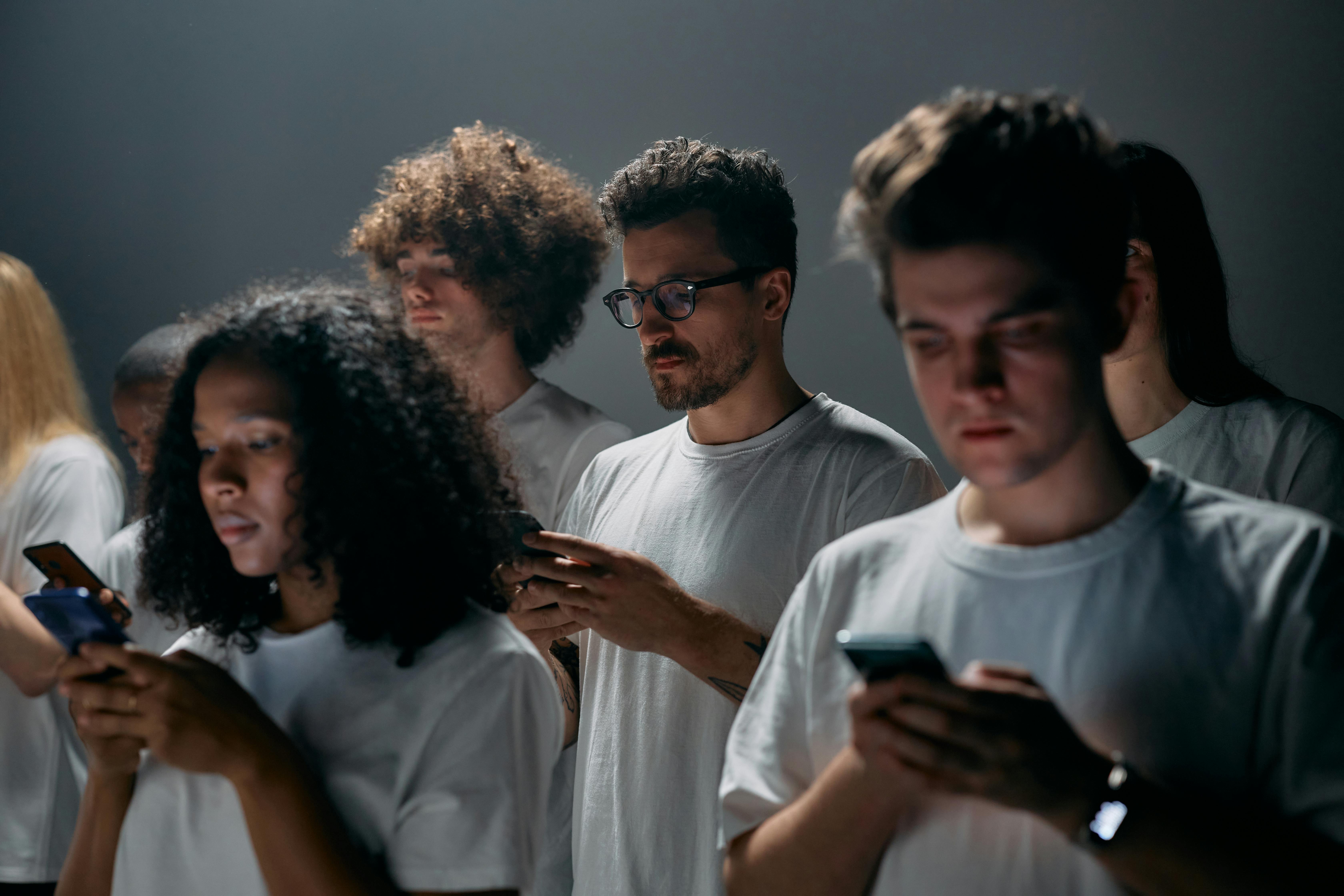 Photograph showing a group of people wearing white shirts, each focused on their smartphones. The setting features dim lighting with a neutral background, emphasizing individual engagement with mobile devices.