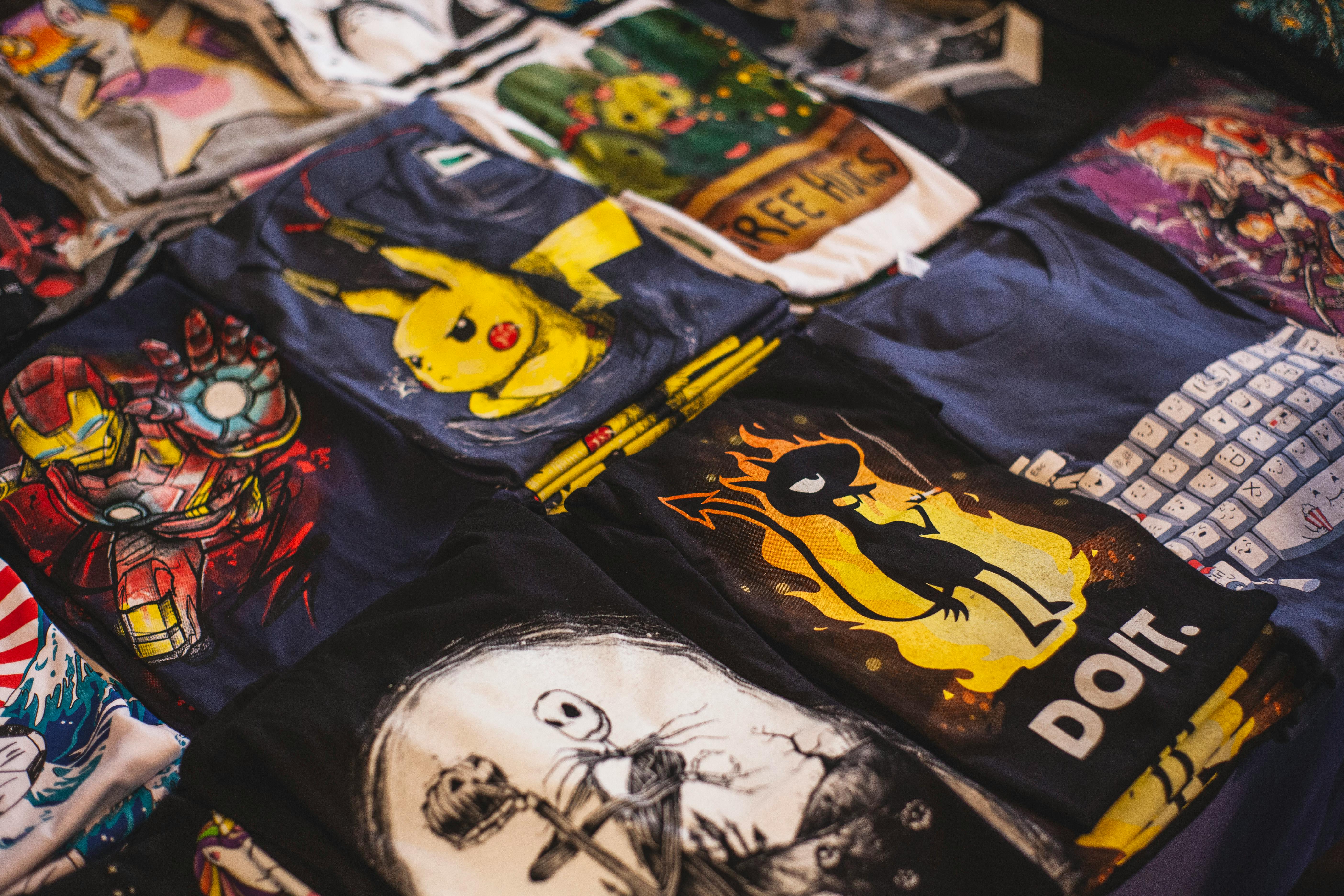 Photograph of various graphic t-shirts laid out on a flat surface, featuring popular culture designs like Pikachu, Iron Man, and a stylized figure with flames and the text "DO IT." The shirts display vibrant colors and distinct artwork, highlighting themes from cartoons, comics, and motivational graphics.