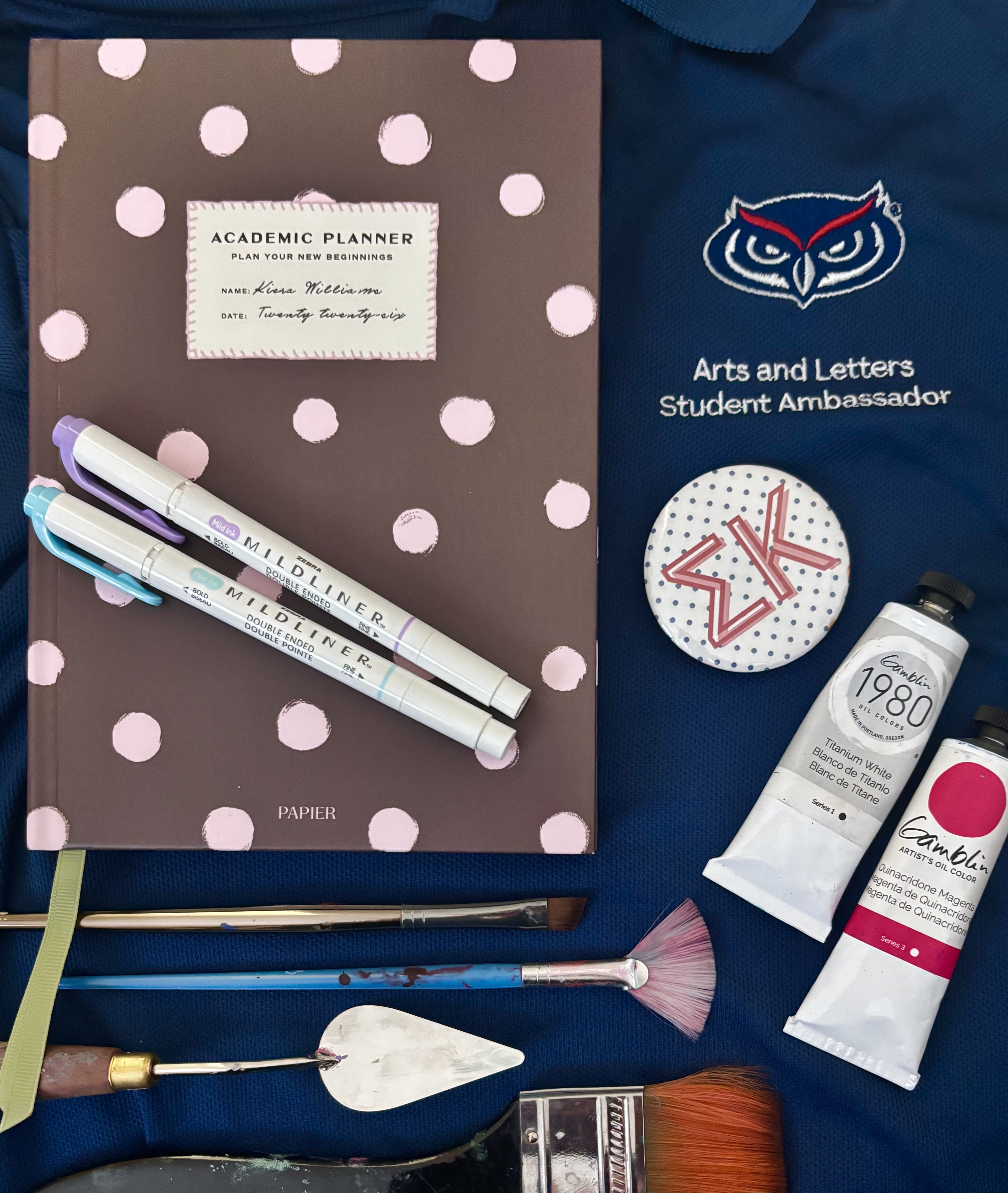 A blue fabric pouch with an owl logo and “Arts and Letters Student Ambassador,” beside a brown polka-dot academic planner, pens and brushes, a small paint tube, and a round badge with a red “4K” marker.
