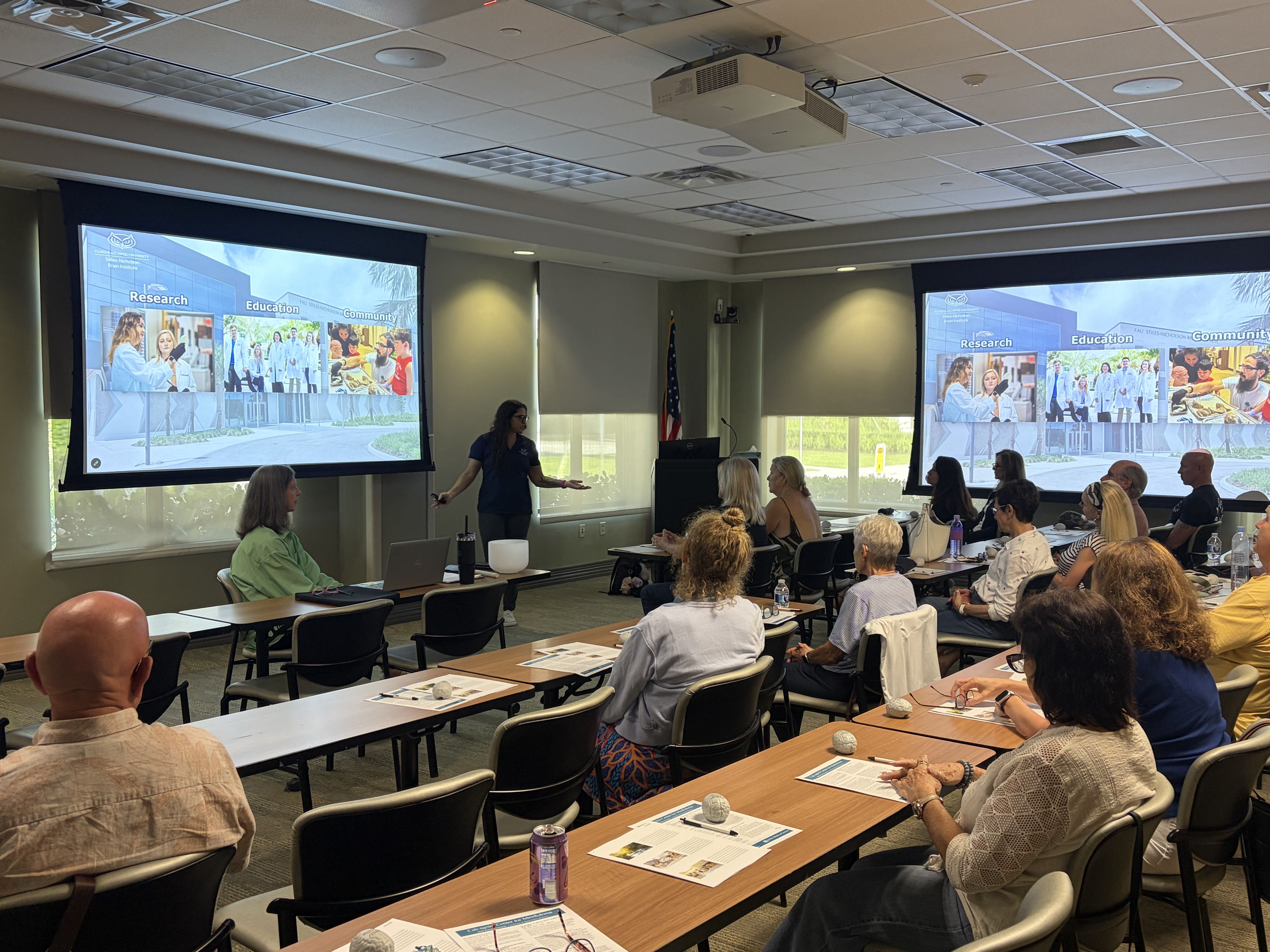 2 projector screens with 3 images (one of a scientist looking in a microscope, one with a group of young scientists walking together, and one with a father and his young son looking at a sheep brain). 3 long tables have people seated facing the screens.