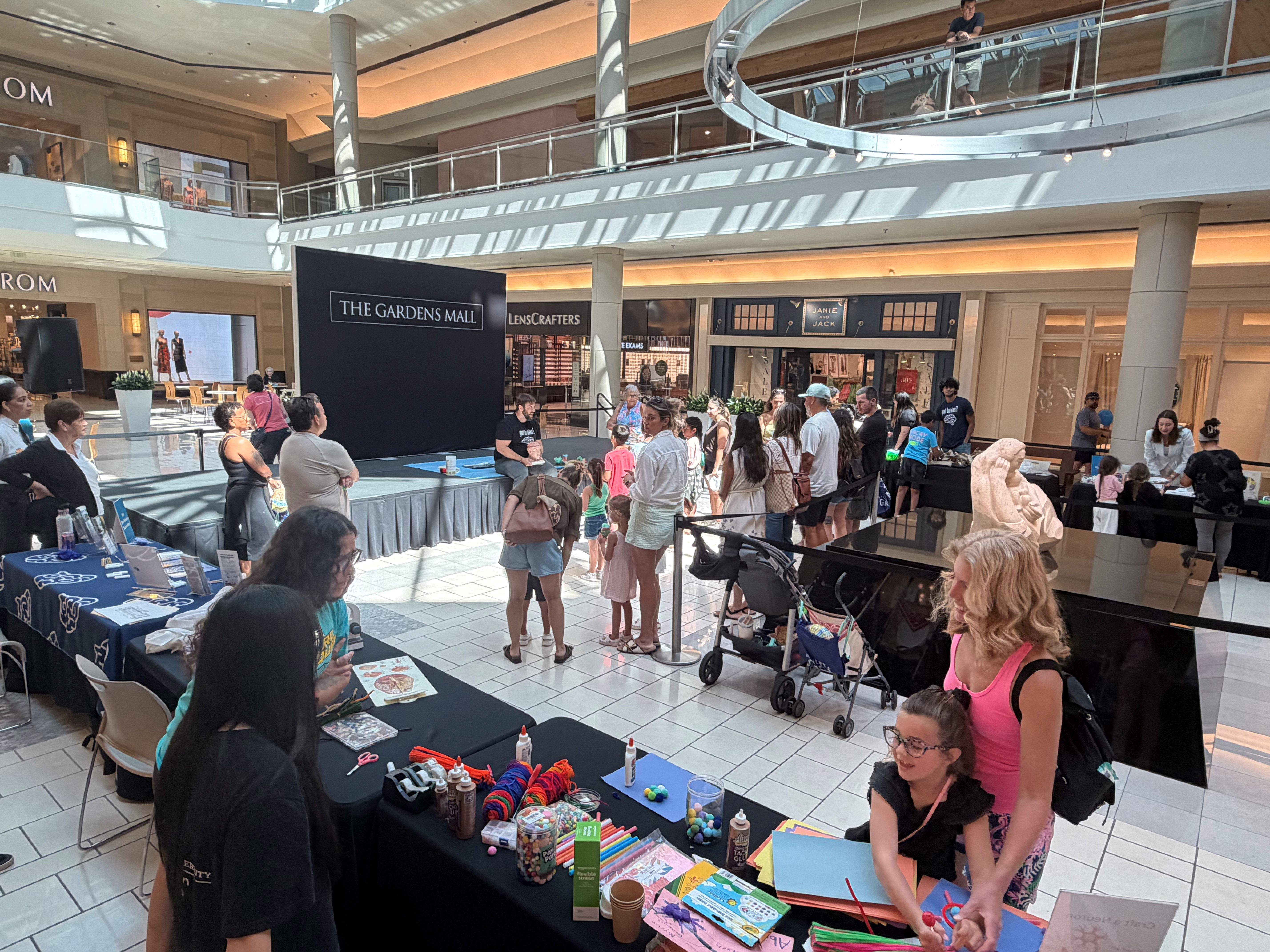 Large stage with a sign that says 'The Gardens Mall" above it. Tables with craft materials and children.