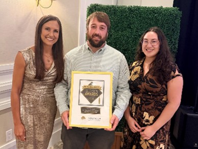 3 people standing in a line facing the camera. the person in the middle is in the center, with a woman on either side of him. they are wearing business formal attire and the man is holding an award certificate