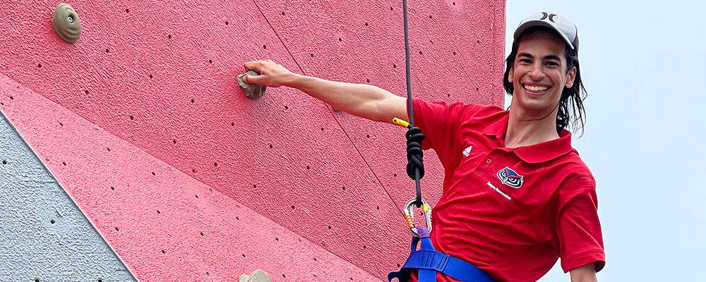 A student smiling as he hangs from the climbing wall