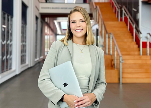 A young, professionally-dressed woman holding an Apple laptop while standing in front of you