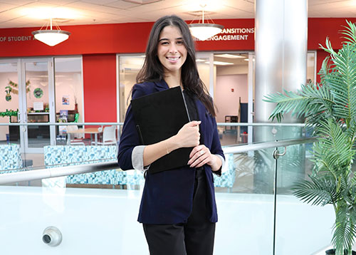 A young, professionally-dressed woman holding an Apple laptop while standing in front of you