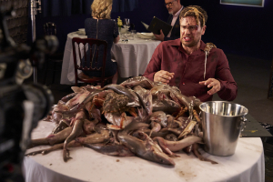 A man in a maroon button-down shirt sitting at a dining table covered in a large pile of fresh seafood, reacting with distress while being filmed on a set.