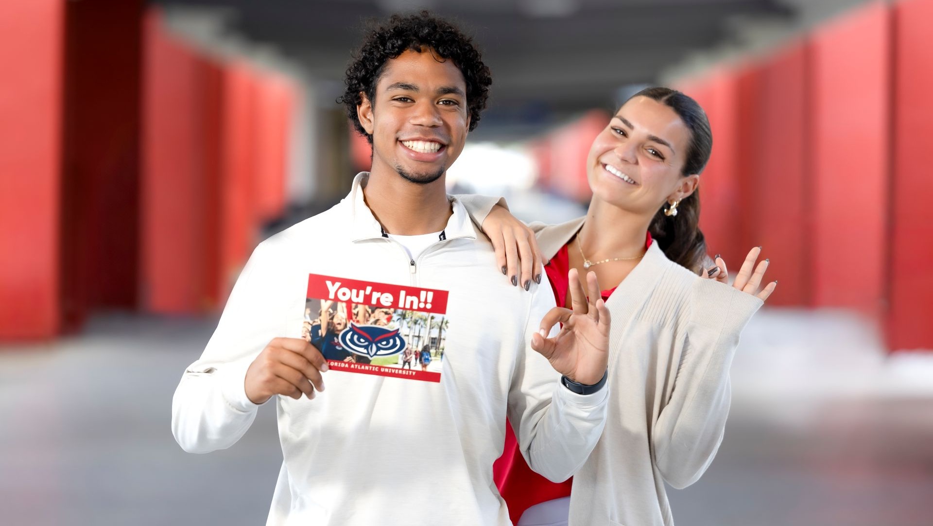 Two smiling individuals standing in a corridor with red pillars, one holding a Florida Atlantic University acceptance card that reads 'You're In!!' with cheering students pictured on it.