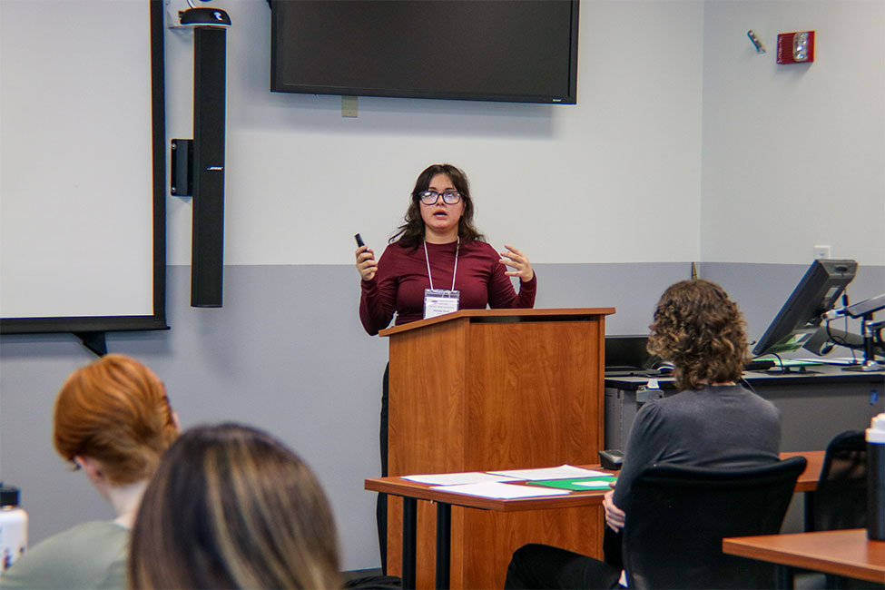 Individual standing behind a podium in a classroom setting addressing people seated in the room. The person is wearing glasses and gesturing while speaking.