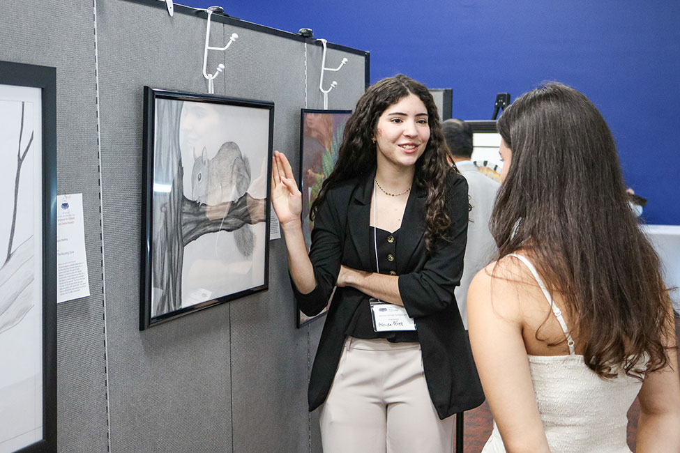 A person stands next to a framed drawing of a squirrel that is displayed on a temporary wall in a room with other artwork on display. The person has a hand on the picture frame and is speaking to another individual whose back is to the camera.