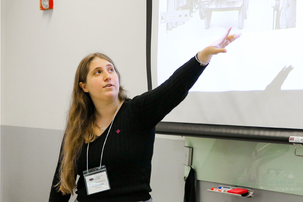 Person stands in front of a projector screen where a presentation slide is displayed. The person is gesturing up to the something on the screen that is not visible.