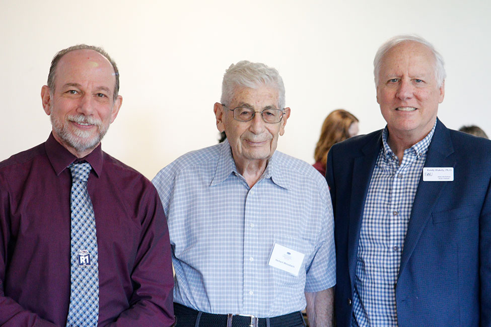 Three people stand side-by-side smiling at the camera in an indoor setting.