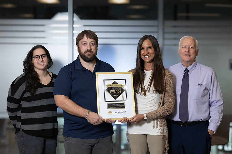 Four individuals stand side-by-side in front of a glass wall. They are all smiling at the camera. The two individuals in the center of the line are holding a framed award plaque.