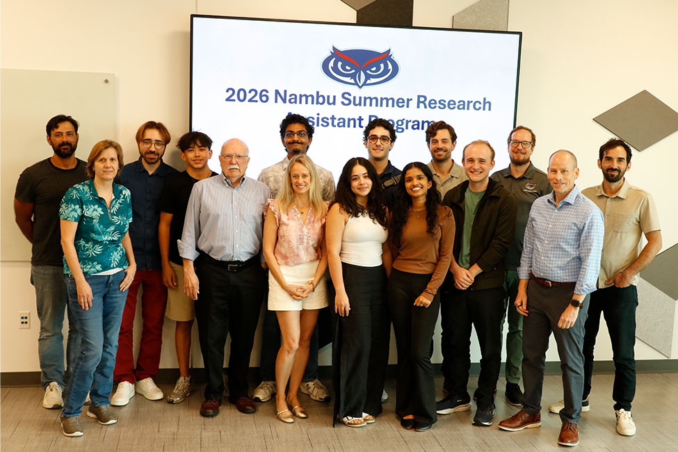 Group photo of about 15 people standing in two rows in an indoor room with wood-look flooring. Behind them is a large screen displaying an owl logo and the text "2026 Nambu Summer Research Assistant Program." The group includes a mix of men and women in business-casual attire, smiling at the camera.