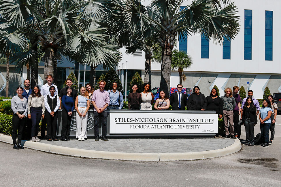 Group photo of students and faculty standing outdoors in front of the Stiles-Nicholson Brain Institute building at Florida Atlantic University. The diverse group poses behind a large campus sign reading “STILES-NICHOLSON BRAIN INSTITUTE – FLORIDA ATLANTIC UNIVERSITY,” surrounded by tall palm trees and modern white architecture under a sunny blue sky.