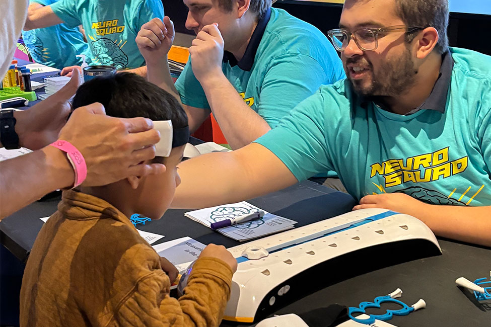 A young child sits at a table while an adult places a small adhesive pad on the side of the child’s head. Across from them, a smiling man wearing glasses and a teal “Neuro Squad” T-shirt engages with the child. Another person in a similar shirt sits nearby. The table holds papers, markers, and a device with buttons, suggesting a hands-on science or educational activity.