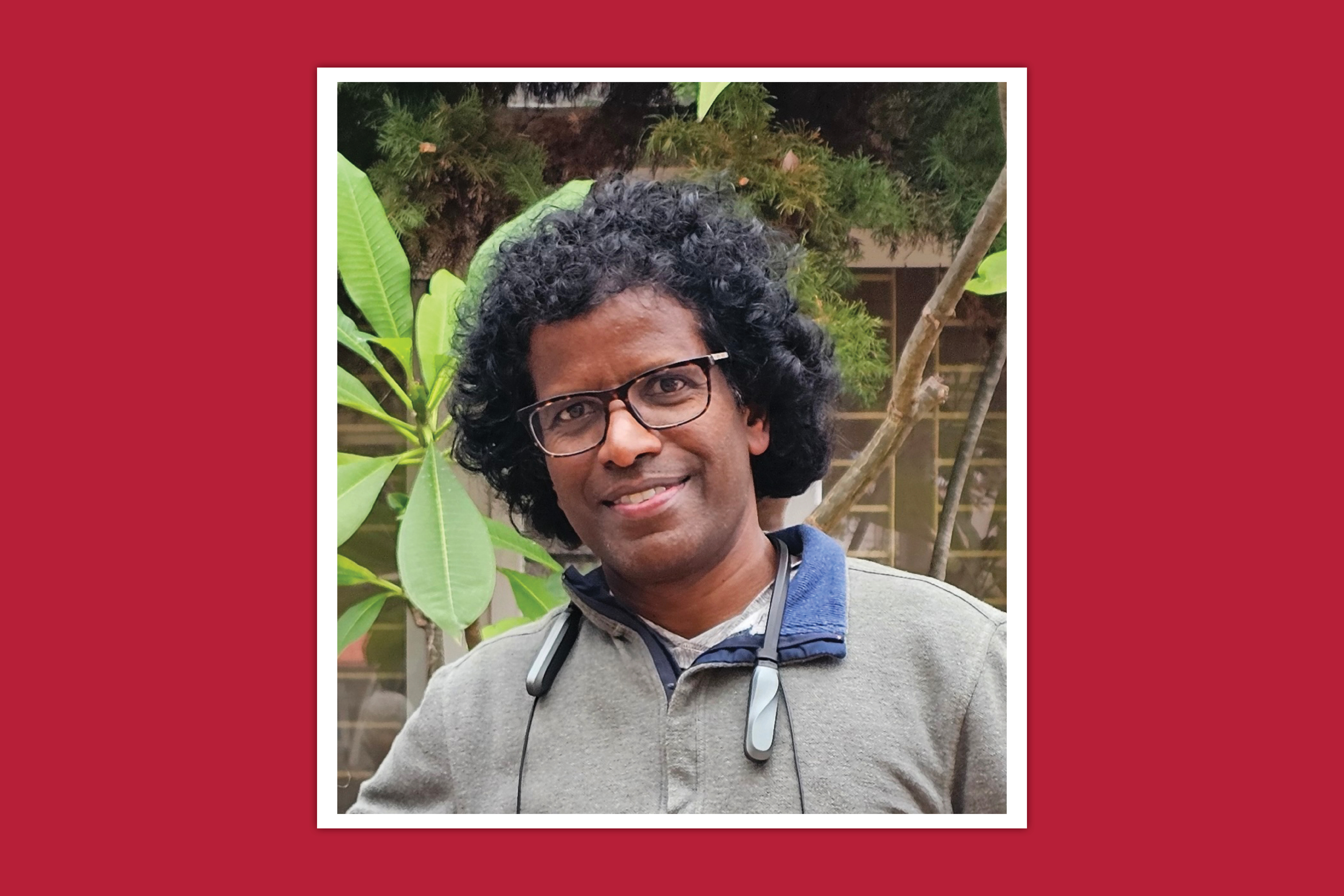 Headshot of a person standing in an outdoor setting with plants and a brick wall in the background.