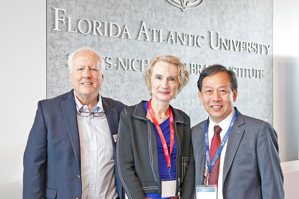 Three individuals stand in a row smiling at the camera with a gray wall behind that reads "Stiles-Nicholson Brain Institute".