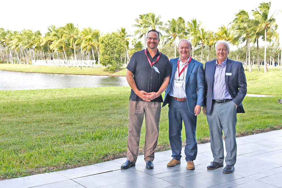 Three individuals stand posing for the camera in an outdoor setting on a sunny with palm trees, a retention pond, and a sign for Florida Atlantic University in the background.