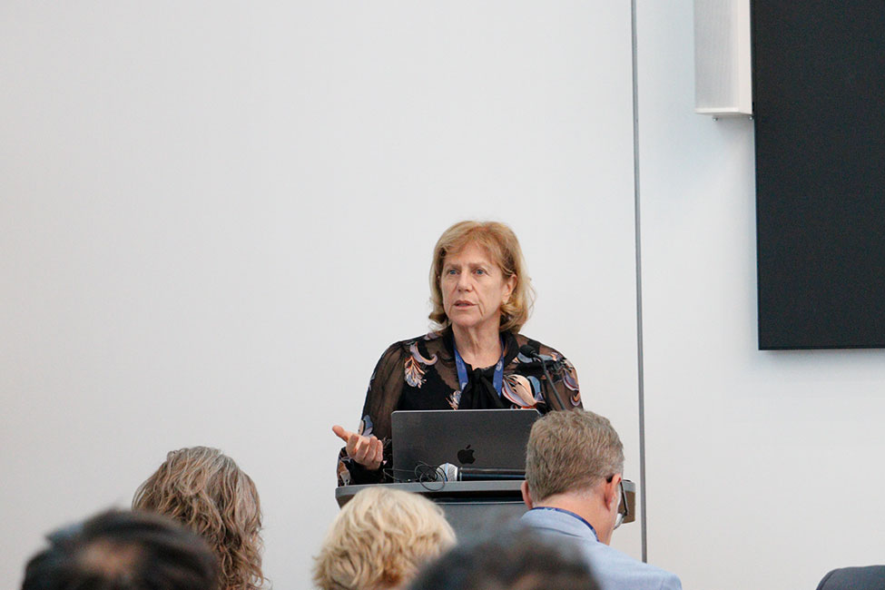 A speaker stands behind a podium talking to a seated audience in an auditorium setting.