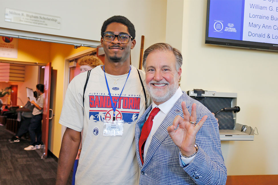 Two individuals posing for a photo indoors, one wearing a "Florida Sandstorm" Florida Atlantic university T-shirt, and the other in a patterned blazer making an FAU Owl sign with their hand.