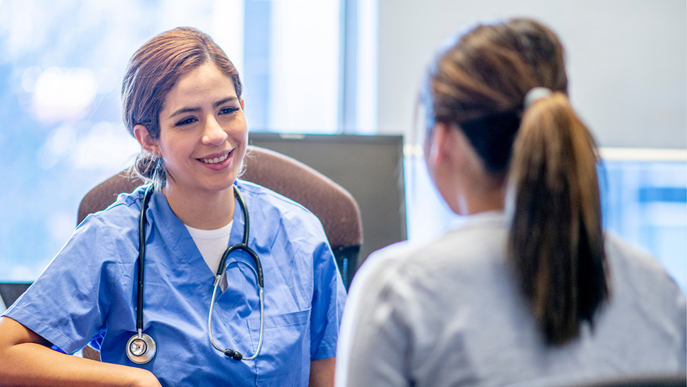 A healthcare professional wearing blue scrubs and a stethoscope sits in a clinical office, speaking with a patient whose back is facing the camera across a desk.