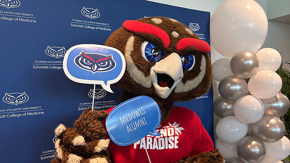 Owlsley, FAU Mascot holding a MedOwls Alumni and Owl logo sign at COM Alumni Reunion