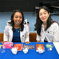 Two medical students in white coats sit behind a blue table displaying food products and clear cups filled with cotton balls representing different levels of fat content. Food items on the table include a box of graham crackers, a Hershey's chocolate bar, and a container of sour cream, each paired with a cup containing a different number of cotton balls to visually demonstrate the fat content in each product.
