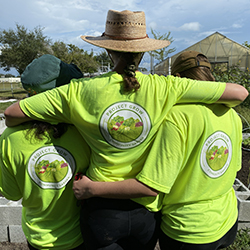 Three people stand side by side with their arms around each other, facing away from the camera. They are wearing bright green shirts with a round "Project Grow" logo on the back. The group is outdoors, near a garden with raised beds and a greenhouse visible in the background, under a partly cloudy sky.