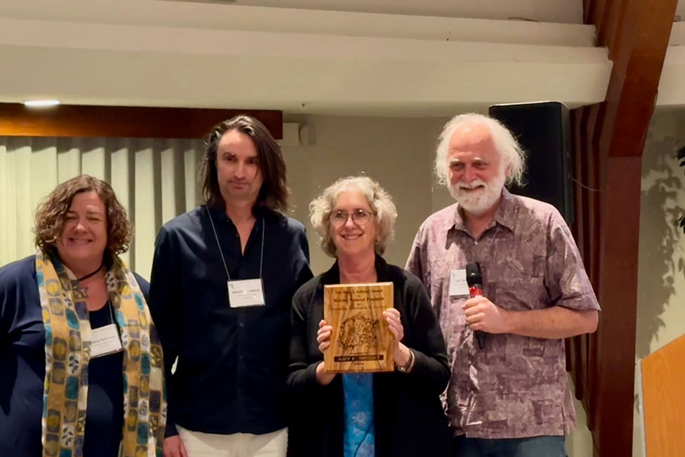 Four people standing side-by-side smiling. The third person from the left, a woman, is holding a wooden award plaque.