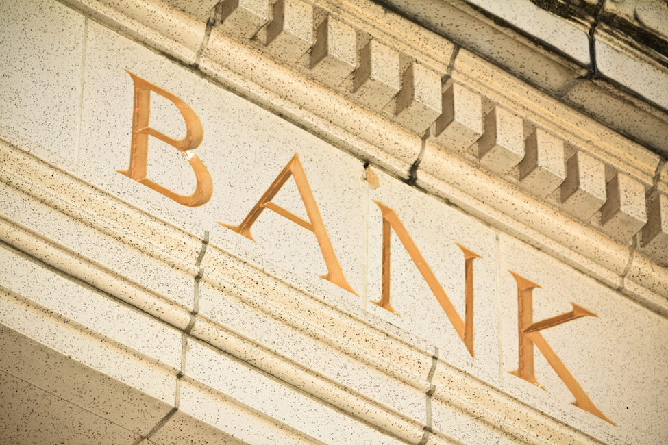 A photo of the top of a stone beige colored building with the word "bank" in all capital letters.