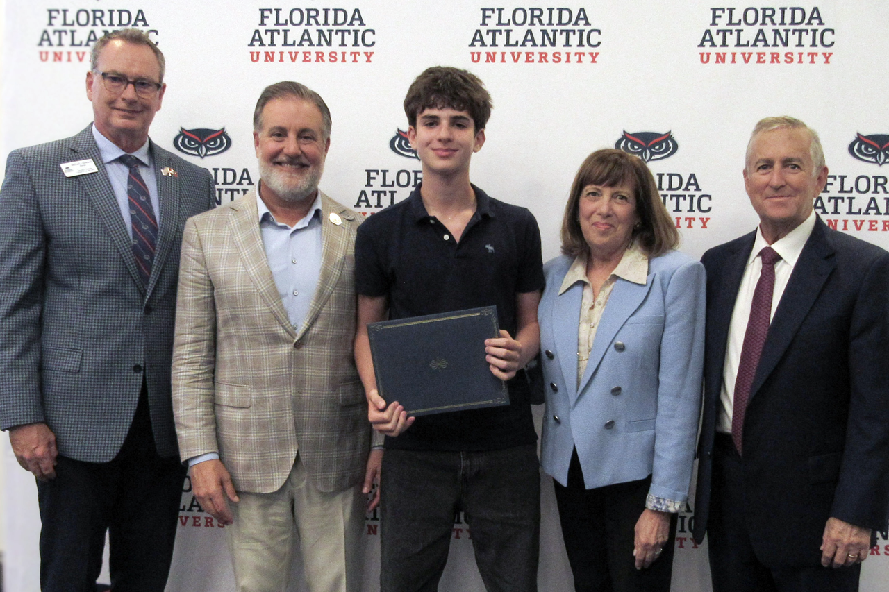 Five individuals stand in front of a backdrop with "Florida Atlantic University" logos. The person in the center holds a certificate or award.