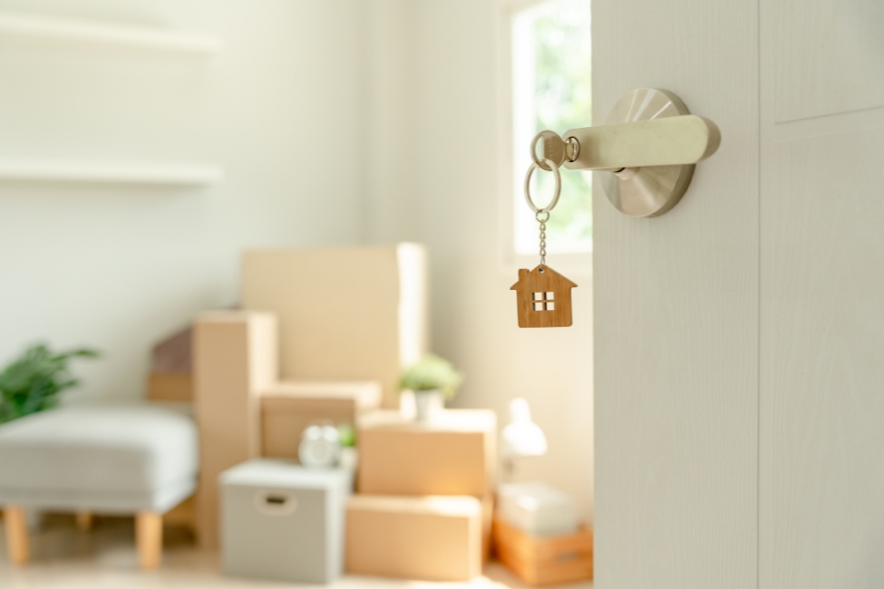 A photo of a white door with a silver handle with a small house key. In the background is a collection of furniture and moving boxes. 