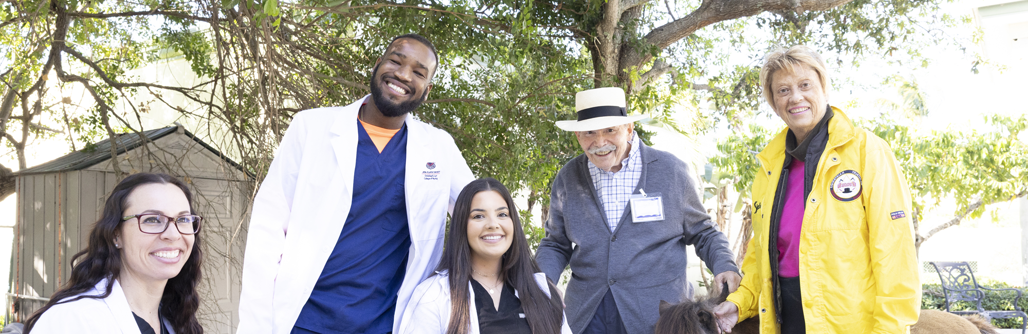 Two individuals in dark blue scrubs smiling outdoors with green foliage in the background.