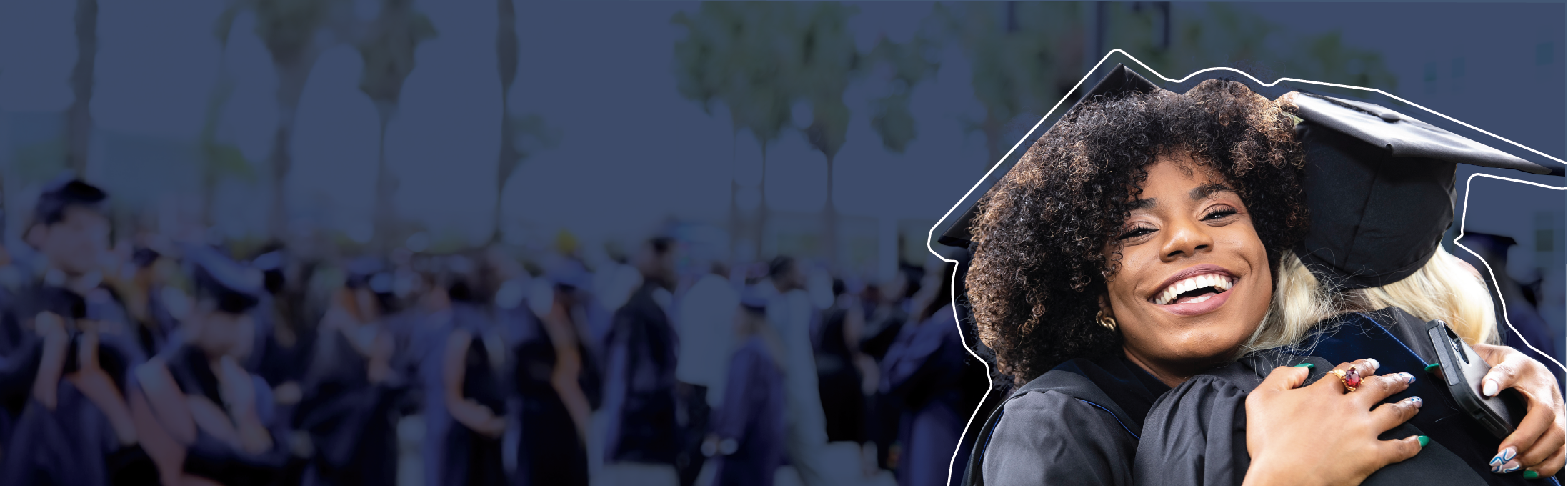 A student in a graduation cap and gown at commencement smiling and hugging another student.