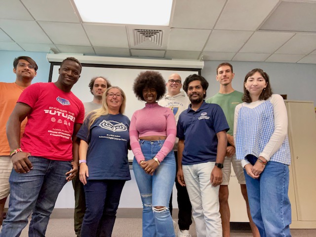 A group of people standing together in a classroom in front of a projector screen, posing for a group photo. Several are wearing shirts with logos, and the room has a tiled ceiling and light-colored walls.