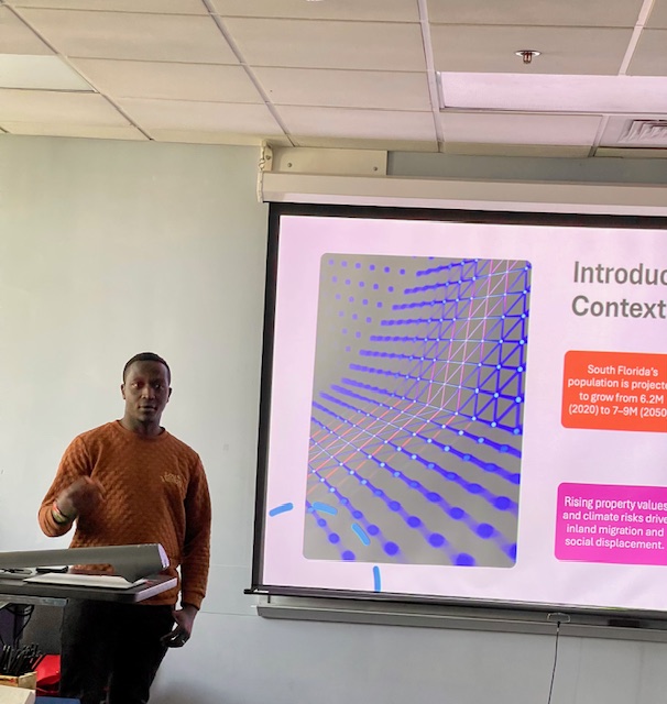 A person presenting next to a projected slide discussing “Introduction and Context,” featuring a graphic of a 3‑D grid and text about population projections and climate‑related impacts.